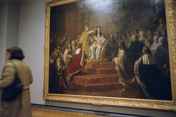Color film photograph (1985) of a 17th-century oil painting of King James II’s coronation hanging in a museum gallery; off-center composition with the gilded frame slightly cropped on the right; English Baroque workshop style with thick impasto, visible craquelure, and warm ambered varnish; rich reds and golds on the dais and regalia; soft track lighting causing a mild glare on the varnish; neutral gray wall and parquet floor; a blurred museum visitor mid-step at the far left edge; no labels or wall text visible; shot on Ektachrome 100 with fine grain and a slight blue cast; 50mm lens on a Nikon F3, handheld, subtle barrel distortion; small dust motes in the air.