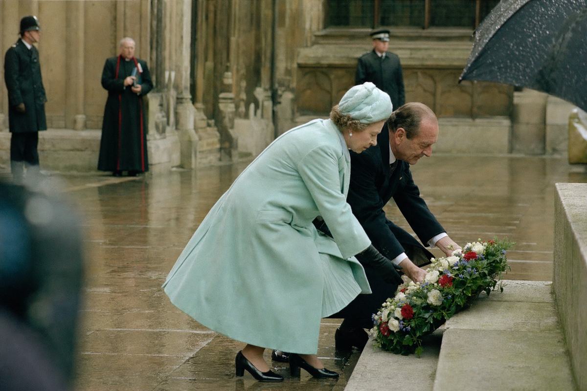 Color press photograph (1985) taken at Westminster Abbey’s cloister during the tricentennial ceremony: the British female sovereign and the French President both mid-bend as they lay a wreath at a stone plinth; oblique composition with the figures to the right, damp paving stones reflecting soft overcast light; distinct faces and natural posture, a cleric and two security officers blurred in the background; no visible text or ribbons readable; shot on Fujicolor 400 with a slight green cast and moderate grain; 85mm lens, handheld, faint motion blur; umbrellas and rain speckles at the frame edge.