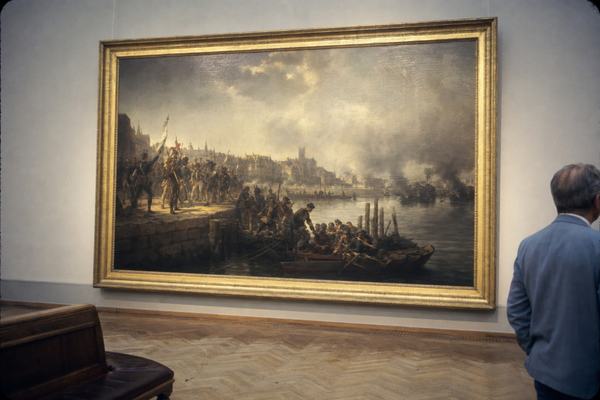 1971 color photograph inside a European museum gallery showing a large 19th-century academic oil painting of Jacques Cathelineau’s entry into Nantes: smoke over the Loire, militias along the quays, boats clustered by timber pilings. The canvas hangs slightly off-center on a neutral wall in a gilded wood frame; parquet floor with scuffs, a bench at an angle, and a single middle-aged visitor partially cropped at the edge. Shot on 35mm Ektachrome with a 50mm lens; natural film grain, slight cyan cast in shadows, gentle vignetting at corners, faint reflection on varnish, no labels or wall text visible.
