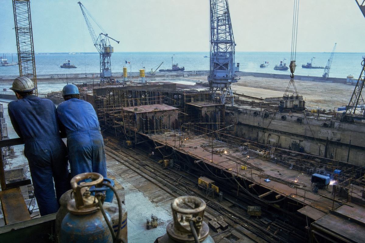 1969 color photograph of the Saint-Nazaire shipyards: full basins with a large merchant hull under construction, lattice cranes, and welding arcs flickering. Royal ensigns flutter on small service craft out in the roadstead beyond the breakwater. A pair of workers in overalls and scuffed hard hats stand near oxy-acetylene tanks at frame left, backs turned. Shot on 35mm Ektachrome with a 28mm wide-angle; slight barrel distortion, salt spray on lens edge, high-contrast steel surfaces, no visible signage or ship names.