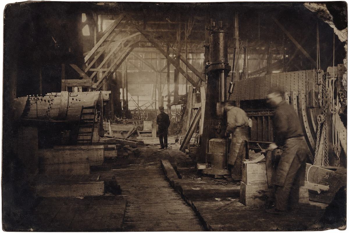 Circa 1865 wet-plate collodion photograph (albumen print from a glass negative) of the Penhoët workshops: an interior view across a slipway with timber blocks, a steam hammer and riveted frames. Long exposure yields slight motion blur on two workers in leather aprons; lantern light pools on oily floorboards. Oblique composition showing rafters and pulley lines; albumen paper sheen, mild sepia tone, corner abrasions, and uneven developer marks near the edges.