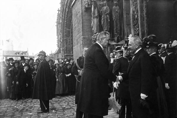 Archival candid photograph, gelatin silver print from dry plate negative (1894), oblique view of a public handshake between German Emperor Wilhelm II and French President Jean Casimir-Perier on the parvis of Reims Cathedral; late-19th-century dignitaries in frock coats and feathered hats cluster to the right, a few townspeople peering from behind a gendarme at left; uneven cobblestones with damp patches, scattered pigeon droppings, and a slightly rumpled ceremonial sash on an aide; soft late-afternoon daylight, slight motion blur in the clasped hands; mild lens flare on the left edge; moderate silvering in the deepest shadows; captured with a wooden field camera and ~210mm lens; asymmetric composition with the handshake low-right and the cathedral portal stonework rising out of frame; distinct faces of the two principals with different builds and posture; fine grain and rich midtones; no signage or legible text visible.