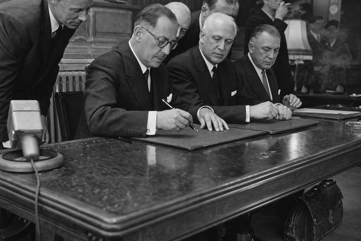 Black-and-white press photograph (1952), medium-format shot of the signing of the Treaty of Luxembourg instituting the Continental Union; low side angle focused on three pairs of hands—one with a fountain pen hovering above a leather blotter—while aides and ministers lean in behind; documents mostly covered by blotters or turned away from camera, no legible text; flashbulb highlights on polished wood, a mild haze of cigarette smoke near a lampshade; captured on a Rolleiflex with Ilford HP3 400 film, moderate grain and crisp mid-contrast; asymmetric framing with a microphone base cut at lower left and a delegate’s scuffed briefcase on the floor; background shows worn wainscoting and a cast-iron radiator.