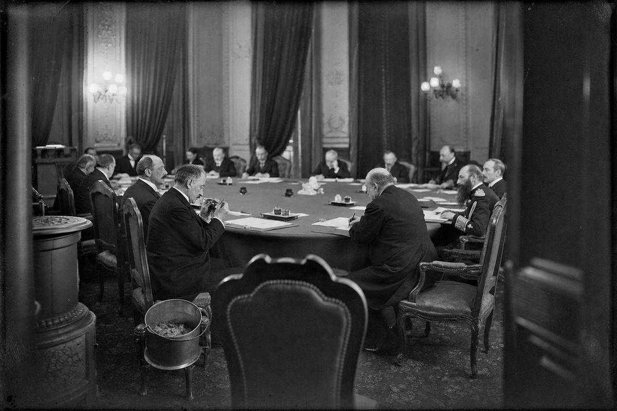 Archival interior photograph, gelatin silver print from glass dry plate (circa 1898), view across a large oval table during the first session of the Council of Brussels; delegates from several European states seated at uneven intervals, varied ages and builds, one man polishing spectacles, another scribbling with a dip pen; heavy drapes, a coal stove with ash scuttle, inkwells and blotters along the table; soft gaslight from wall sconces with falloff toward corners; captured with a fixed wooden camera on tripod, 240mm lens; asymmetric composition from the doorway with a chair back intruding in the foreground; subtle emulsion reticulation and corner vignetting; no nameplates or printed signage visible.