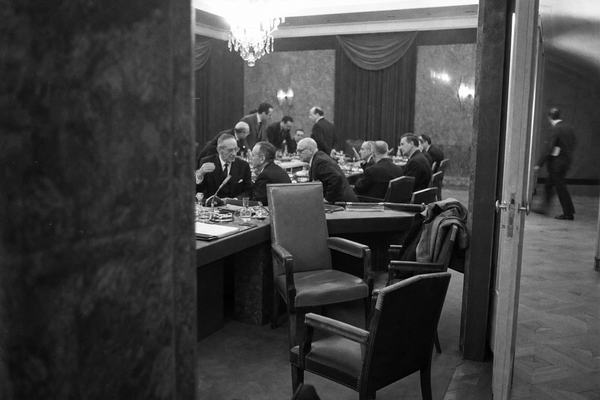 Archival black-and-white press photograph taken in Brussels, 1965, inside the Council meeting room during the Empty Chair period. Oblique viewpoint from behind a pillar onto a long conference table strewn with microphones, ashtrays, water carafes, and folders; one prominently empty chair at a place of honor while delegates from West Germany, Italy, Belgium, the Netherlands, and Luxembourg confer in low voices. Mid-century chandeliers and heavy curtains in background; a coat draped over a chair back; a messenger hurrying past in the corridor beyond a half-open door. Shot on Kodak Tri-X 400 pushed to 800 with a Leica M3 and 35mm lens at f/2.8; pronounced film grain, slight motion blur on a gesturing hand, mild vignetting, uneven exposure near ceiling lights. Asymmetric, candid composition. No visible text, signage, nameplates, or lettering anywhere in frame.