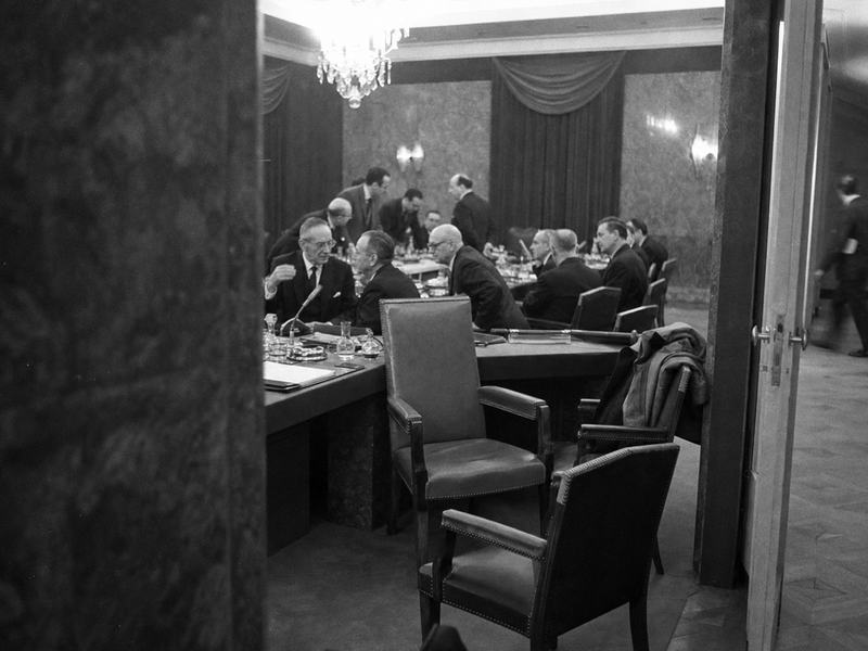 Archival black-and-white press photograph taken in Brussels, 1965, inside the Council meeting room during the Empty Chair period. Oblique viewpoint from behind a pillar onto a long conference table strewn with microphones, ashtrays, water carafes, and folders; one prominently empty chair at a place of honor while delegates from West Germany, Italy, Belgium, the Netherlands, and Luxembourg confer in low voices. Mid-century chandeliers and heavy curtains in background; a coat draped over a chair back; a messenger hurrying past in the corridor beyond a half-open door. Shot on Kodak Tri-X 400 pushed to 800 with a Leica M3 and 35mm lens at f/2.8; pronounced film grain, slight motion blur on a gesturing hand, mild vignetting, uneven exposure near ceiling lights. Asymmetric, candid composition. No visible text, signage, nameplates, or lettering anywhere in frame.