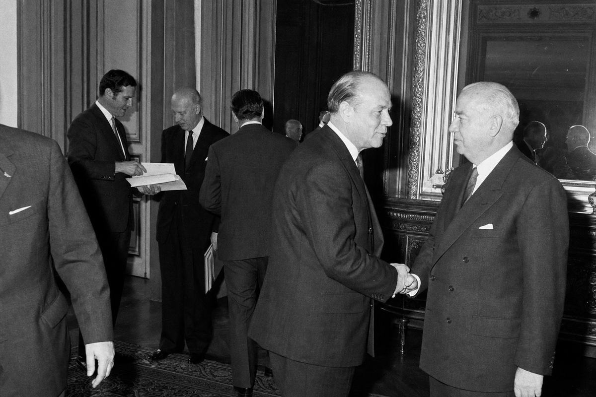 Black-and-white press photograph captured in June 1966 inside the Egmont Palace, Brussels, moments after the Treaty of Brussels signing. Side-corridor scene with wood-paneled walls and a gilded mirror catching reflections; two senior statesmen of the Five mid-handshake, slightly off-center, while three aides of distinct ages and builds cluster in the background comparing notes. A flashbulb has just fired, casting hard shadows and slight overexposure along one edge; visible Tri-X grain. Shot on a Nikon F with a 50mm lens, handheld; asymmetric, candid framing, one figure half-cropped entering frame. Papers clutched but turned away from camera. No visible text, signage, badges, or lettering anywhere in frame.