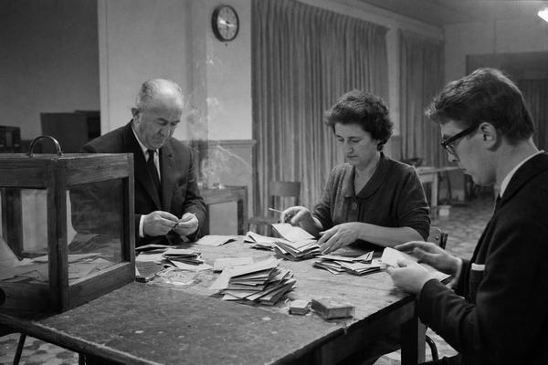 Black and white 35mm press photograph on Kodak Tri-X 400, pushed to 800, taken inside a French municipal vote-counting room late at night on April 27, 1969. Asymmetric composition with a wooden table in foreground, a sealed glass-and-wood ballot box to the left, and three distinct election officials—one older man in a dark suit with thinning hair, a middle-aged woman in a modest dress with rolled sleeves, and a younger man with horn-rimmed glasses—counting folded ballots by hand. Harsh overhead fluorescent lighting, slight motion blur in fingers, cigarette smoke haze drifting across frame, wall clock partly visible indicating late hour. Scuffed linoleum floor, chipped paint along baseboards, heavy curtains in background, no visible text or logos anywhere. Shot with a 50mm lens, natural film grain and uneven exposure at edges.