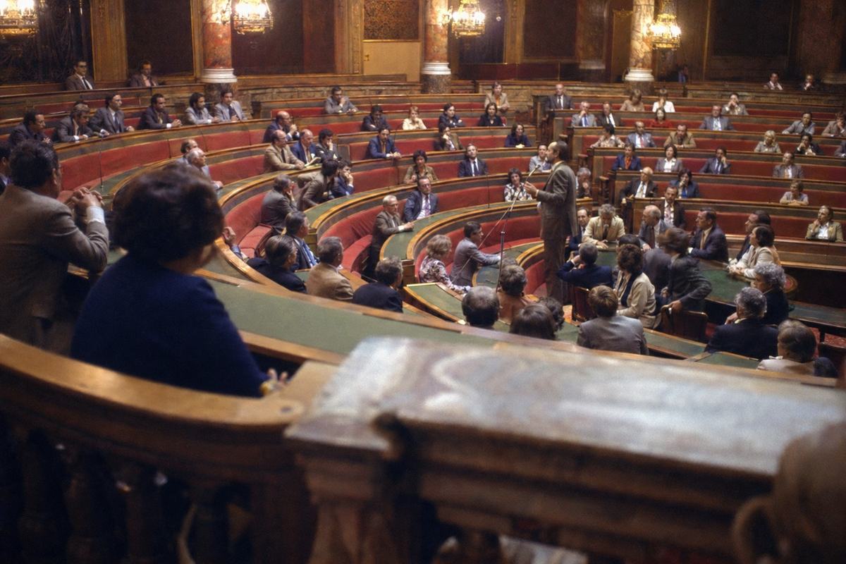 1970s color photograph on Kodak Ektachrome 64, slight tungsten warmth and mild magenta cast, wide-angle (28mm) view from the public gallery of the hemicycle at the Palais du Luxembourg during a session of the newly installed territorial chamber. Asymmetric frame: the foreground balustrade soft-focused, the curved rows of red-upholstered benches and green leather-topped desks below. Delegates—men and women of varied ages—leaning in to listen; one figure standing at a microphone mid-speech. No papers spread on desks, no nameplates, and no signage or text visible. Warm chandeliers create gentle halation; subtle motion blur on one turning head; fine Ektachrome grain, shallow depth of field falls off into the back rows.