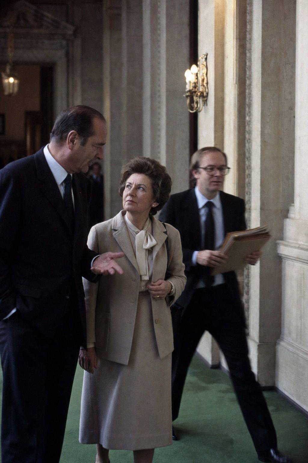 Late-1970s color photograph on Kodak Ektachrome 200, slight cyan cast, candid corridor scene inside the Palais du Luxembourg outside a committee room. Vertical framing with an off-center trio: a tall regional president in a dark suit gesturing with an open hand, a shorter woman in a beige skirt suit listening intently, and a bespectacled aide holding a stack of plain, unmarked folders. Stone columns and worn green carpet visible, brass wall sconce flaring slightly; no signage, no door plaques, no visible text anywhere. Taken with a 85mm lens; subtle motion blur in the aide’s step; period-accurate grain and color shift.