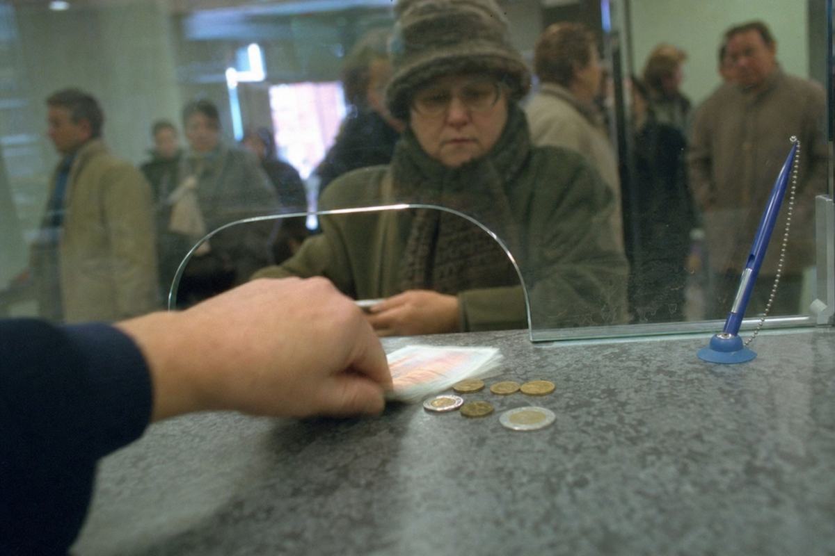 Color 35mm photograph, late 1999, bank counter in Calais; teller’s bare hand slides the first ‘Union’ banknotes and a handful of coins across a scuffed laminate counter; shallow depth of field so denominations and lettering are out of focus (no legible text); Fujicolor Superia 400 on Nikon F100 with 35mm lens; cool fluorescent cast with slight green tint; mixed-age customers queue blurred in background, winter coats, plastic pen chained to counter, acrylic shield with smudges; slight motion blur on the passing note; asymmetrical, close framing.