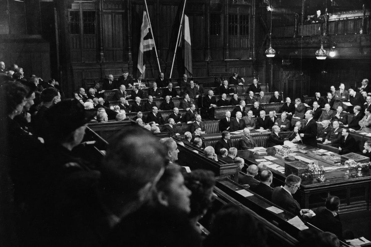 Black-and-white silver-gelatin photograph, 1948, inaugural joint sitting of the Assemblée de l’Union at Westminster; vantage from the public gallery, oblique angle across mixed benches of MPs and députés; dual flags on poles at the far end, partially obscured; members leaning together to confer, one member mid-speech, papers spread on green benches; Speed Graphic 4x5 with 127mm lens; Kodak Super-XX sheet film, pronounced grain in shadows, slight lens falloff, flashbulb highlights on varnished wood; dust motes in light shafts; asymmetrical composition; no legible text.