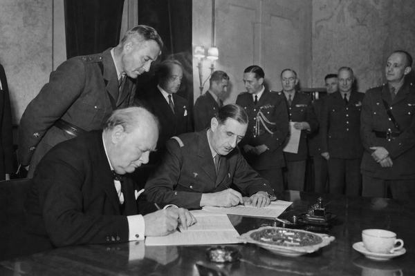 Archival silver-gelatin press photograph, June 27, 1940, interior of 10 Downing Street; candid over-the-shoulder view as Winston Churchill and General Charles de Gaulle lean over a polished table signing a joint union act; British and French staff officers of varied ages and builds stand behind at uneven distances; ashtray with stubbed cigars, scattered papers, tea cup ring stains; wartime blackout curtains, dim tungsten lamps with slight smoke haze; Leica IIIa with 50mm lens, handheld; panchromatic film on nitrate base, medium grain, slight motion blur on Churchill’s hand, uneven exposure near the lamp; off-center composition; background wall maps partially out of frame; no legible text anywhere.