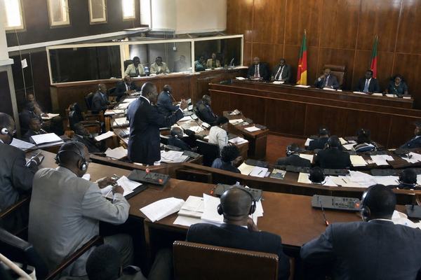Digital press photograph, early 2000s, inside the Federal Senate chamber in Buea during a plenary session; shot from the upper public gallery with a 24mm wide-angle lens, oblique composition capturing wooden desks with microphones and scattered papers, senators in formal attire with simultaneous-interpretation headsets, discreet national flags near the rostrum, interpreter-booth glass catching faint reflections; warm mixed lighting from high windows, slight high-ISO noise and mild motion blur on gesturing hands, uneven exposure across the room, natural skin texture and distinct faces at varied ages and builds; documentary newsroom style, candid, no banners or signage in frame.