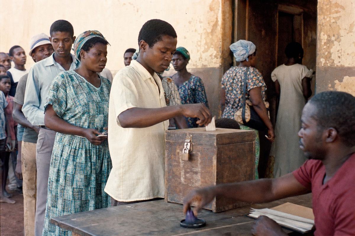 Color photograph on Kodachrome 64, May 20, 1972, polling place in Buea; 50mm lens at eye level, asymmetrical line of voters inching toward a scuffed wooden ballot box with a metal latch on a rough table; sunlit dust in the air, warm color cast, visible film grain and subtle edge vignetting; slight motion blur on an official’s hand reaching for an ink pad; natural skin texture, varied ages and builds; background earthen wall with peeling paint and a shaded doorway; press-documentary style, candid, no legible signage.