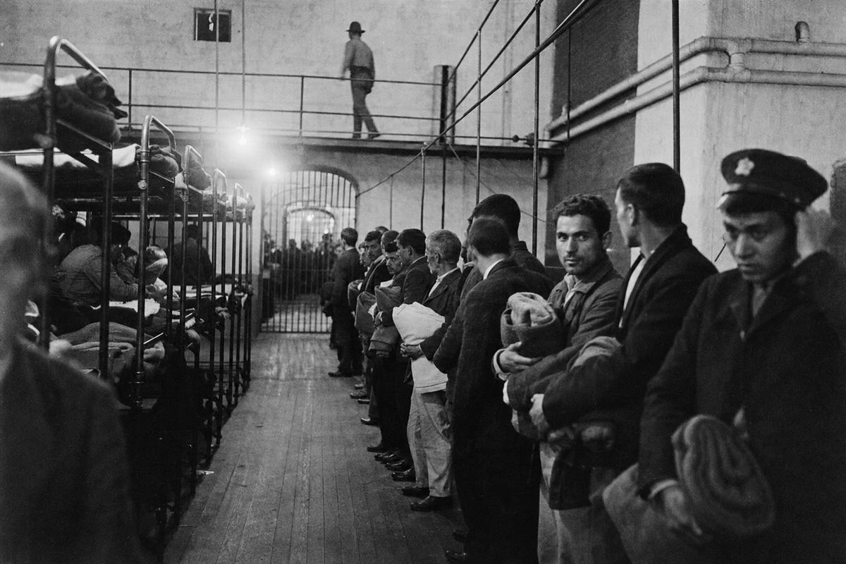 Archival silver gelatin print from the early 1920s inside the Ellis Island detention annex: oblique view down a barracks corridor with rows of iron cots; a guard in a campaign hat walks the catwalk above while a line of internees with folded blankets waits below; distinct faces of varied ages and builds, some weary, one man adjusting his cap; scuffed floorboards, steam pipes along the wall, bare bulbs casting halation; photographed with a 4x5 press camera on orthochromatic film; medium grain, foreground slightly out of focus, edge vignetting; dominant gesture is the man looking toward a gate at the far end; no legible signage.