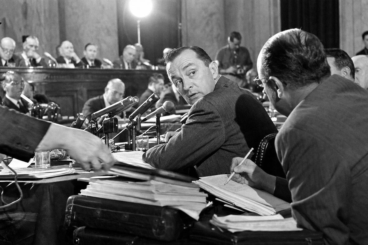 Mid‑1950s press photograph inside a Senate Internal Security Committee hearing room: low three‑quarter view from the press row showing a witness at a crowded counsel table under hot lamps, wired microphones stretching toward him; counsel leans in with a pencil mid‑note while an aide’s hand blurs past a stack of folders; senators sit out of focus on a raised dais in the background; shot on Tri‑X 400 with a Speed Graphic and flashbulb; high contrast, specular highlights on microphones, slight edge vignetting; dominant moment is the witness turning his head toward an unseen questioner; no legible lettering on nameplates or documents.
