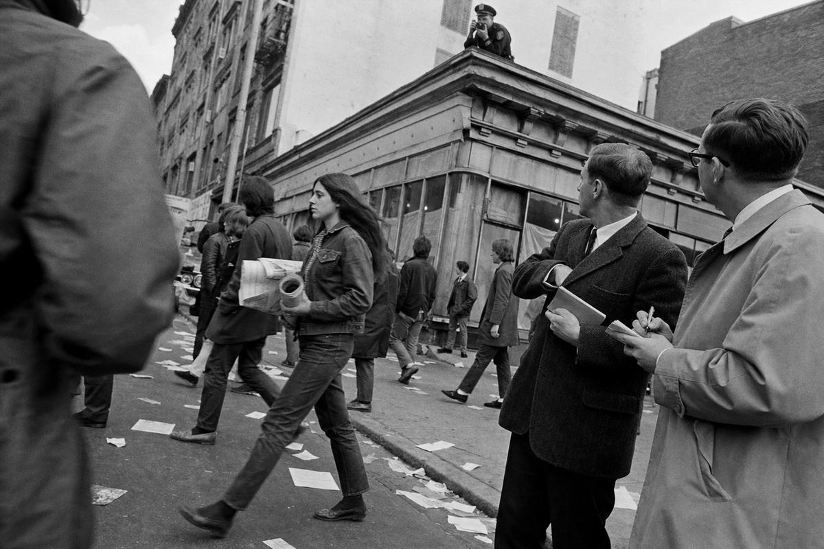 Late‑1960s street protest captured on 35mm Tri‑X 400: oblique street‑level view of marchers passing a storefront; a young woman in a denim jacket strides mid‑frame carrying a rolled banner, while two plainclothes men linger at the curb taking notes and glancing toward a rooftop where a uniformed photographer peers over a parapet; scattered leaflets trampled underfoot with no legible text; pronounced grain, high contrast, slight barrel distortion from a 28mm lens, 1/125 shutter with motion blur in legs; dominant gesture is a plainclothes observer tucking a notebook into his coat.