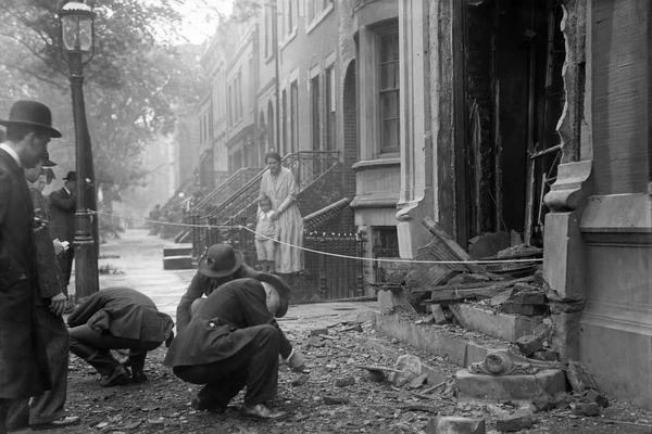 Archival silver gelatin photograph taken at dawn on June 3, 1919, showing the blasted front of Attorney General A. Mitchell Palmer’s R Street, Washington, D.C. residence; detectives in brimmed hats stoop to examine debris on the scorched stoop while a neighbor in a housecoat steadies a child just behind a rope line; splintered doorframe and bricks scattered across the sidewalk, a gas streetlamp and adjoining rowhouses receding into soft morning haze; photographed on a 5x7 view camera with an uncoated lens, dry plate negative; moderate grain, slight motion blur on a detective’s hand, uneven exposure at frame edges; asymmetric composition with the torn doorway off to the right and figures clustered low left; no signage or legible text visible.