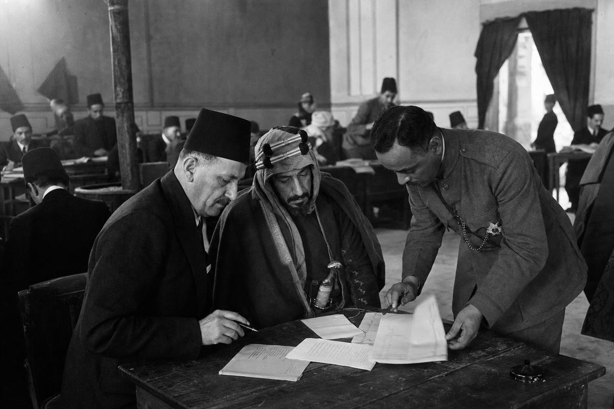 Archival silver-gelatin photograph from late 1918 inside a Damascus assembly hall during the Damascus Charter deliberations; 4x5 press camera with magnesium flash, slight smoke haze visible in overhead light, shallow depth of field from a 127mm lens. Candid, off-axis framing: three delegates in varied dress—one in a dark frock coat with a fez, one in tribal cloak with a silver-hilted dagger at his belt, and one in khaki with Sharifian insignia—lean over a scuffed wooden table strewn with annotated pages and an inkwell, while the larger hall recedes in soft focus with benches, a coal stove, and a curtain pulled back to a side corridor. Uneven exposure at the edges, subtle motion blur on a turning page; faces distinct and unposed.