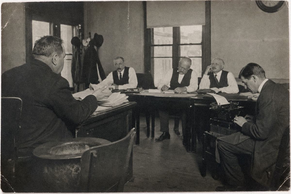 Circa‑1905 arbitration hearing: counsel addresses a three‑person panel as a stenographer types in a sunlit room.