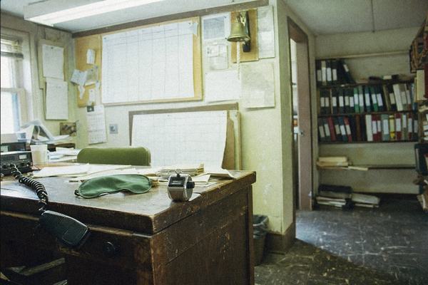 35mm color photograph on Fujicolor Superia 400, taken with a 35mm lens from shoulder height inside a small 1990s office at 238 Bank Street, New London. Asymmetric composition: a cluttered oak desk sits left foreground with a green eyeshade and a metal mechanical tally counter; pinboards fill the midground with taped lane-draw grids and paper weigh-in sheets, a small brass bell mounted above. Shelves of binders with colored spine stripes line the right wall; fluorescent ceiling tubes cast a slight green cast while morning window light reveals floating dust motes. Noticeable 400-speed grain, slight corner softening, and mild fluorescent flicker; shallow depth of field keeps any paperwork and binder markings out of focus, no readable text anywhere. Floor scuffed, a coiled VHF radio mic half off the desk, a cracked baseboard near the door. No watermarks or lettering.