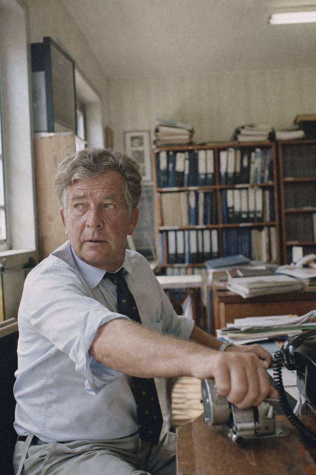 Deputy secretary Martin Keating at his desk, mid-reach toward a tally counter, binders and office shelves behind him.