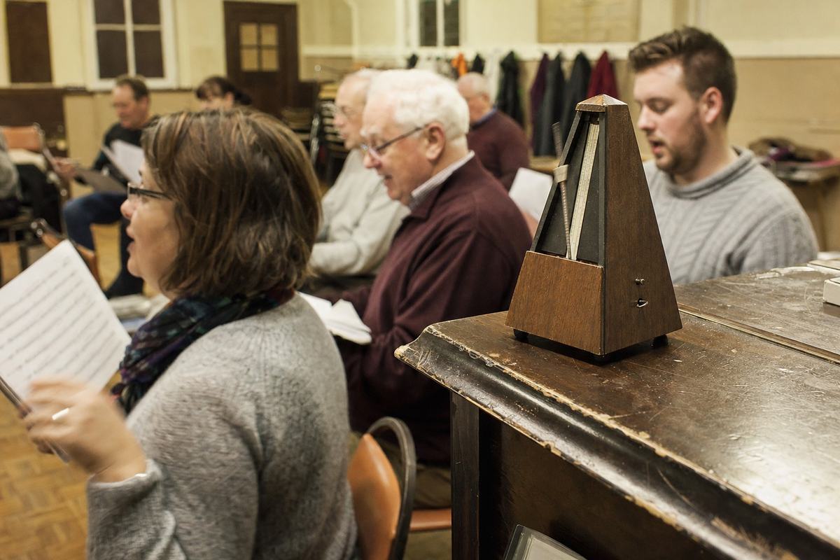 Répétition de chœur dans une salle paroissiale à Barr, un métronome en bois posé sur un piano, des choristes feuilletent leurs partitions.