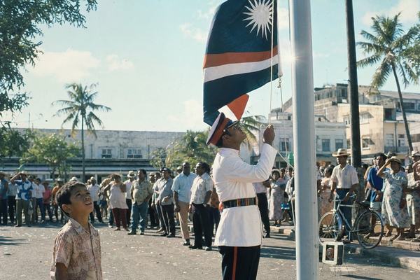 1972 color photograph, street-level documentary photojournalism in a tropical city center during a sovereignty ceremony; shot on a 35mm Nikon F with a 50mm lens using Fujicolor 100 film; slight cyan-green cast, gentle contrast, visible film grain and minor edge softness; asymmetric composition with the dominant subject being a uniformed guard hoisting a new national flag on a rope while a young child in the foreground looks up, mouth slightly open; varied islander crowd behind them in period-accurate clothing (short-sleeve shirts, summer dresses, straw hats), natural skin texture, imperfect posture; palm trees and low-rise stucco buildings in the background, humid air shimmer, scattered confetti on asphalt; no readable text, labels, or signage anywhere; candid expressions; incidental details like a bicycle leaning against a post and a portable speaker on the curb; balanced exposure under midday sun with a hint of lens flare from the flagpole