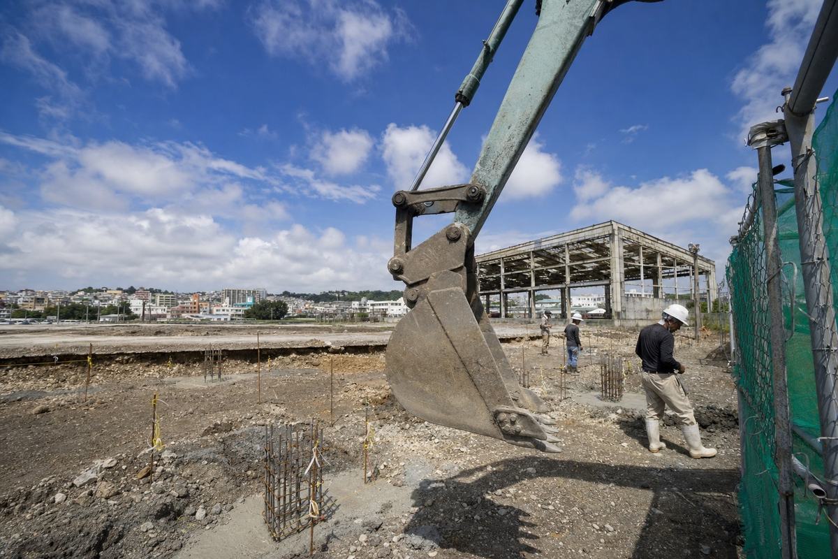 Contemporary digital press photograph inside the former MCAS Futenma footprint during early redevelopment works in 2022; shot with a 24mm wide-angle lens, midday glare with polarized sky; dominant subject is an excavator’s bucket paused above marked ground stakes and rebar cages; in the midground, the skeletal frame of an old hangar and concrete pads; Ginowan’s low skyline and a patchwork of rooftops beyond; chain-link fencing with green mesh partially frames the right edge; no visible signage, logos, or lettering; workers in hard hats moving naturally, distinct ages and builds; asymmetric composition with the excavator arm cutting diagonally across the scene