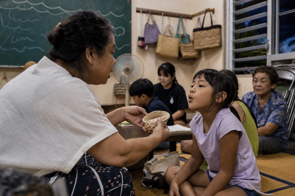 Contemporary digital press photograph inside a modest island community center during an evening Uchinaaguchi language class; shot with a 35mm prime on a mirrorless camera, natural window light, ISO 1600 with visible but fine-grained noise; dominant subject is a middle-aged teacher’s hands holding a seashell while a primary-school girl watches intently, her lips forming a word; other students and an elderly observer softly out of focus in the background; tatami mats, stackable chairs, and a chalkboard with simple shapes but no writing; handwoven bags on pegs, a wall fan, and open louvers letting in sea breeze; no visible text or lettering anywhere; asymmetric composition from slightly below eye-level