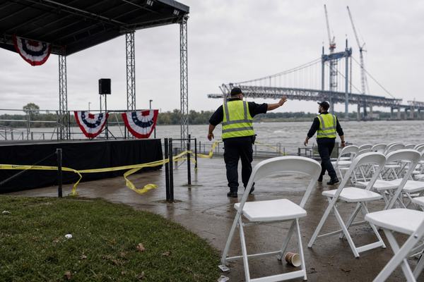 Digital press photograph taken on a cloudy September morning in 2026 at the St. Marshall riverfront park. Asymmetric composition: the empty outdoor stage sits left-of-frame with wind-tossed bunting; foreground right shows rows of unused white folding chairs at slight angles, one chair leg near a discarded paper coffee cup. Yellow security tape flutters across a scuffed concrete walkway. Two security officers in reflective vests, mid-40s and late-20s with distinct faces and builds, move people along in the mid-ground, heads turned away from camera. In the background, partially visible steel spans of the unopened Huron Gate Bridge cross the river, soft focus. Overcast sky, rippled river surface, damp grass patches, light litter at the edge of the path. No visible text, logos, or signage. Shot on a full-frame mirrorless camera with a 35mm lens at f/5.6, 1/500s, ISO 400; natural light; slight wind blur on bunting; subtle noise in shadow areas; documentary news photo style.