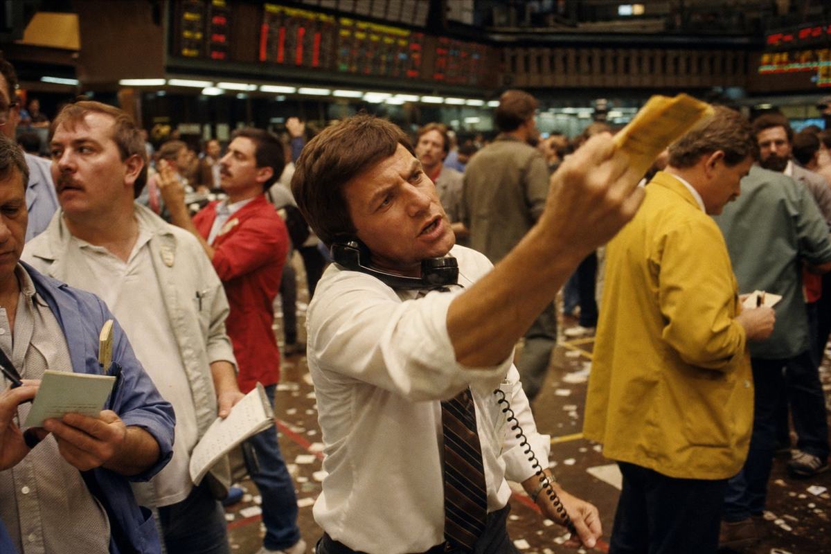 Color photograph from the 1980s taken inside the USFE trading floor in Philadelphia; 35mm SLR with a 28mm lens at shoulder height; Kodachrome 64 film under tungsten ambient light producing a warm amber cast; a trader in a white shirt and striped tie holds a telephone against his shoulder while raising a yellow ticket in his other hand, face strained in concentration; behind him, distinct colleagues of varying ages and builds gesture and scribble on pads; overhead electronic price boards emit a soft raster glow without any readable characters; paper litter on the scuffed floor, taped lane markings partly visible; slight motion blur on hands, fine film grain, uneven exposure at frame edges; asymmetrical, candid composition.