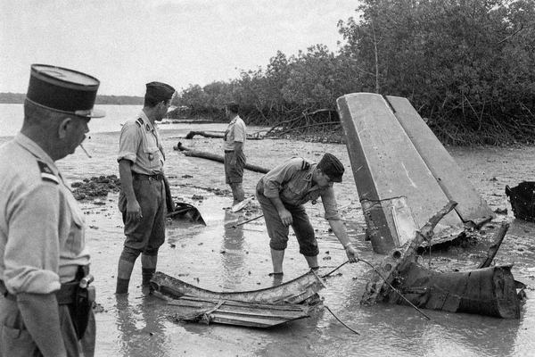 Black-and-white 1950s press photograph taken at low tide on the Catembe mangroves opposite Lourenço Marques, Mozambique: Portuguese gendarmes in mixed ages and builds wade ankle-deep in rippled mud among torn aluminum panels and a pusher aircraft tail section half-sunk and tilted; one gendarme bends to touch a control cable while another, slightly out of focus, watches with a cigarette; tangled mangrove roots and a beached log in the background, overcast sky; mud splashes on uniforms, scuffed leather boots; asymmetric, candid framing with the wreck off-right; shot on Kodak Tri-X 400 with a 50mm lens from a staff reporter’s handheld camera; moderate film grain, slight motion blur on a turning head, uneven exposure at the frame edge; no text, no signage.