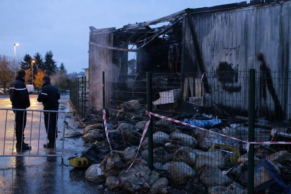 2016 digital press photograph taken at dusk outside the burnt postal sorting center in Brives-Charensac; shot on a Canon 5D Mark III with a 35mm lens at f/2.8, high ISO noise and slight motion blur from handheld capture; asymmetric composition with the charred hangar and partially collapsed roof beams in midground right; scorched and water-damaged mail sacks spilled on wet asphalt; police tape strung haphazardly across a dented metal fence in the foreground; two gendarmes in dark jackets stand near a temporary barrier at left, distinct faces turned away in candid posture; faint smoke residue streaks on corrugated walls; shallow puddles reflecting blue evening light and a few distant streetlamps; scattered debris like broken plastic crates; cool autumn air visible as breath; no legible text or signage anywhere; straightforward documentary style, slightly uneven exposure at the frame edges.