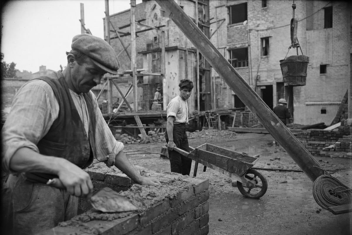 Silver gelatin dry-plate photograph, circa 1926, municipal housing construction site in Paris’s 19th arrondissement. Asymmetric framing: in the near left foreground a mason in a flat cap trowels mortar onto a rising course of brick; behind him a younger laborer with rolled sleeves steadies a wheelbarrow; timber scaffolding and a pulley bucket cut diagonally across the frame. Background shows a half-built courtyard block with raw brick and open window bays; muddy ground with pooled water, scattered planks, and a coil of rope. 9x12 cm plate camera with 135mm lens; orthochromatic sensitivity rendering the sky pale; crisp midtones, fine grain, slight motion blur at the mason’s trowel, mild edge vignetting from the lens hood. No legible signage.