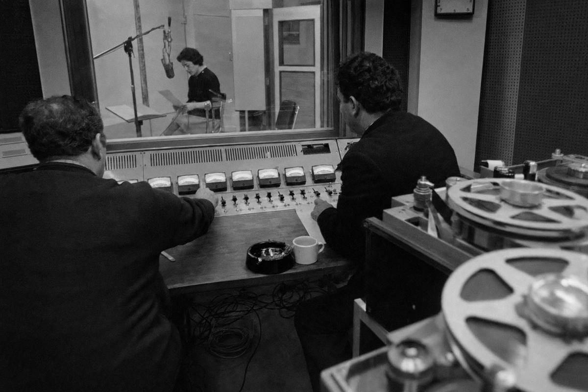 ORTF recording studio, early 1960s, session in progress: press photograph on Kodak Tri-X 400, 35mm lens; two sound engineers at a console with glowing needles, one hand mid-fader; through the window, a seated actress reads into a large dynamic microphone; open-reel tape machines with large spools, ashtray and coffee cup on the edge of the desk, coiled cables on the floor; acoustic panels and a clock with no legible face details; high-contrast grain, slight motion blur in the engineer’s fingers; asymmetric framing with the control room dominating the right; no text or logos visible.