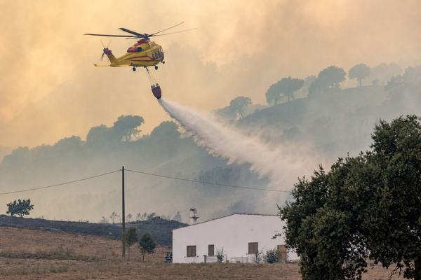 Digital press photograph, 2026, Sardinia near Oristano: a civil protection helicopter with high‑visibility markings (no readable text) releases a water bucket over smoke‑stained cork oak hills; late‑afternoon sun filtered by ash haze; heat shimmer above a ridgeline; a thin power line sags across the mid‑ground, a white farmhouse with closed shutters partly visible beyond a dry field; 70–200mm lens at ~150mm, 1/500s, f/5.6, ISO 800; natural color, slight motion blur on rotor and falling water; asymmetric composition with the helicopter high left and the water arc diagonally toward the right; fine particulate in air catching light; no signage, no watermarks.