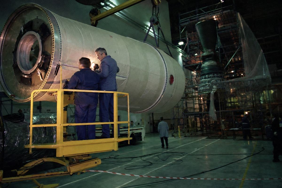 Color photograph on Fujicolor 200, Les Mureaux assembly hall, late 1988: a cylindrical Ariane H stage section hangs from an overhead crane, matte insulation panels catching greenish fluorescent light; two technicians on a yellow scissor lift inspect a seam—one tall with salt-and-pepper hair, one shorter with a heavier build—faces lit by a portable work lamp; floor shows scuffs, coiled cables, and a taped safety line; depth layers of scaffolding and a partially shrouded engine nozzle in the background; shot handheld with a 35mm lens, slight motion blur and pronounced film grain in the shadows; off-center framing with the stage to the left; no text, stenciling, or logos visible.