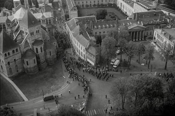 1977 aerial black-and-white photograph taken from a light helicopter over the palatine quarter of Compiègne during the millennial ceremonies; shot on Kodak Tri-X 400 with a Nikon F2 and 35mm f/2 lens at 1/500s; fine-to-medium film grain, slight motion blur at frame edges, gentle atmospheric haze; asymmetrical framing with the Romanesque mass of Saint-Corneille off to the left, the palatial and parliamentary wings to the right; the Place des Plaids below showing a bending civic procession with a brass band turning a corner, scattered spectators, police cordons, and tree-lined ceremonial axes; classic stone facades catching soft spring light; incidental details like parked service vans and litter near a curb; no visible text or signage; realistic documentary style.