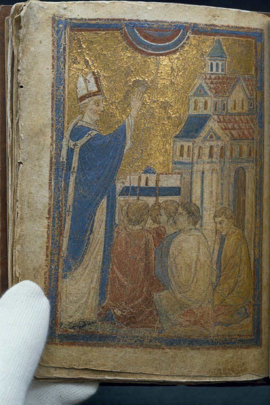 Archival reproduction photograph of a 12th-century Romanesque illumination on vellum from the capitular library of Saint-Corneille, showing the consecration of the church with a bishop’s raised hand and a simplified palatine palace at right; School of northern France, tempera and gold leaf on parchment; visible pigment craquelure, worn gilding, cockled vellum, foxing at the margins; frayed edges and old stitch holes; photographed on a copy stand in 1977 on Ektachrome 64, slight cyan cast, shallow raking light revealing surface relief; cropped tightly to the miniature panel to exclude any inscriptions or lettering; off-center framing with a curator’s gloved hand just entering the lower left to steady the folio; neutral background, no visible text.