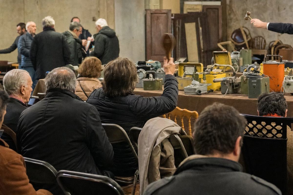 Salle des ventes à Saint‑Ouen: table d’anciens compteurs Geiger et dosimètres, public attentif, une palette se lève.