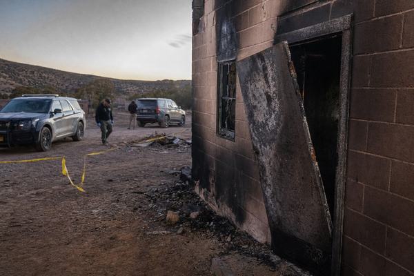 Digital press photograph at dawn outside a rural New Mexico sheriff’s substation; charred cinderblock facade, a burned steel door hanging ajar, bullet-pocked window glass, yellow tape fluttering; two dust-coated patrol SUVs parked at angles, tire ruts and scattered debris in the foreground; pale early light catching airborne ash and dust; distant low hills and scrub visible past the lot; no legible signage; composition from a shoulder-height oblique angle with the door framed in the right third; captured on a full-frame mirrorless camera with a 28mm lens at f/5.6, ISO 3200; visible high-ISO noise in the shadows, slight lens vignetting, and uneven exposure from smoke haze; two investigators blurred in mid-distance, faces indistinct; no text or watermarks.