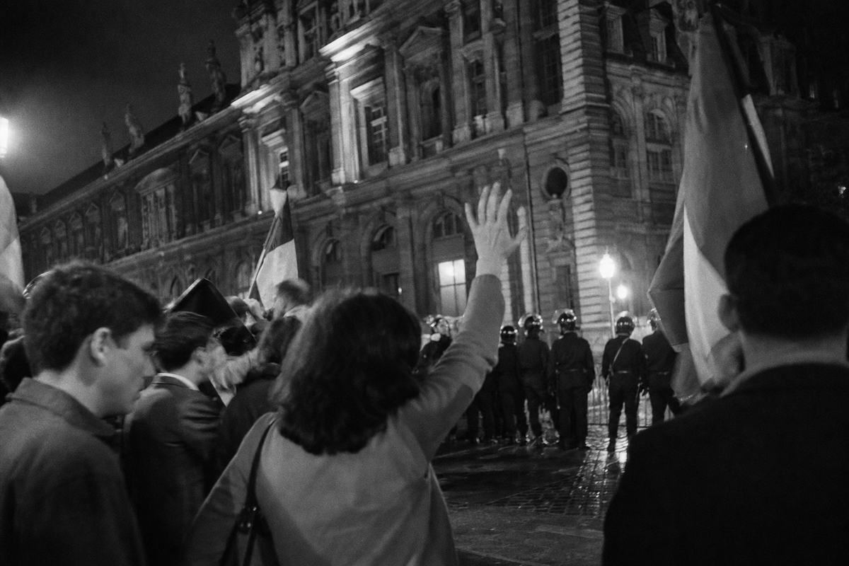 Black-and-white press photograph, Paris, June 1962, evening demonstration on the forecourt of the Hôtel de Ville; low angle from within the crowd with a single woman in foreground raising an open hand, nearby faces blurred in motion; helmeted police line backlit along the curb, wet cobblestones reflecting sodium lamps; tricolor flags partially visible without any lettering; shot handheld on a Leica M3 with 35mm Summicron using Kodak Tri-X 400 pushed to 800; pronounced grain, motion blur on hands, slight flashbulb spill at frame edge; asymmetric composition with the façade receding to the right; no signs or banners with text visible.