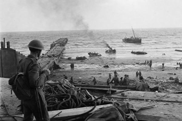 Archival black-and-white silver gelatin photograph, June 3, 1940, Dunkirk beach seen from an elevated perch near the shattered east mole; the broken pier juts diagonally into a grey, wind-chopped sea; smoking wrecks of small boats in the shallows; clusters of exhausted Allied soldiers crouch or move singly on wet sand under a clearing sky; foreground littered with coiled ropes, splintered planks, and overturned tarpaulins; a small trawler listing in the distance; captured with a 35mm Leica III and 35mm Elmar lens on Agfa Isopan film; pronounced grain, slight motion blur from spray, salt-speckled lens, and uneven exposure across the frame from haze; candid, off-center composition typical of 1940s frontline reportage; no text or signage visible.