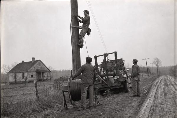 1930s documentary photograph, silver gelatin print on fiber paper, captured with a 4x5 Speed Graphic press camera using a 135mm lens from shoulder height beside a rural dirt road. A WPA-era pole crew installs multi-pair telephone cable onto newly set wooden poles; one lineman on climbing spikes guides the cable while another on the ground braces the reel. Overcast sky, damp soil with tire ruts, barbed-wire fence and a weathered farmhouse soft in the background. Period-accurate work truck parked askew; any decals or placards turned away so no legible text. Asymmetric, candid composition with the worker up the pole as the dominant subject. Material details: slow panchromatic film look, moderate grain, slight motion blur in the lineman’s hands, mild edge vignetting, shallow depth of field characteristic of large-format press work. No readable text or logos anywhere in frame.