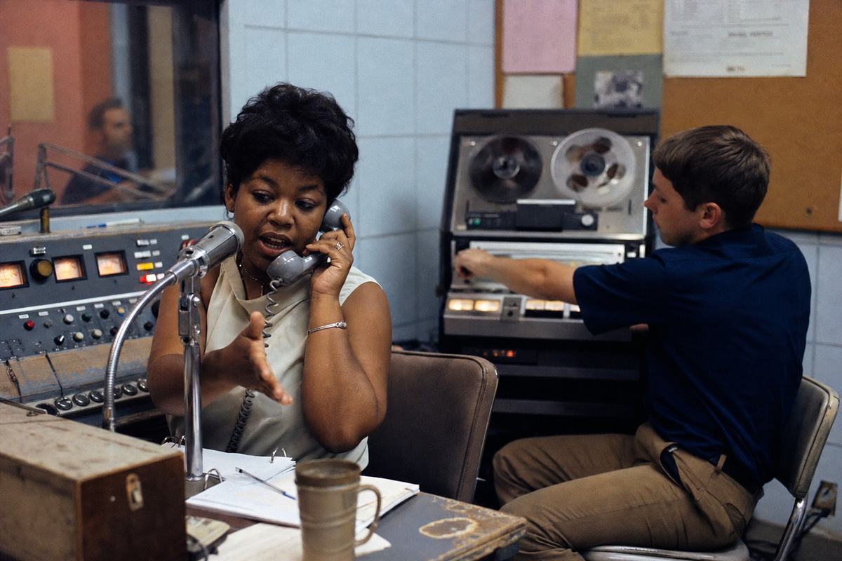 Mid-1960s color photograph on Kodachrome transparency, shot with a Nikon F and 50mm lens from just outside the console desk. Inside a small community radio studio during a live call-in: a Black female host in her 30s leans toward a chrome microphone while cradling a telephone handset; a white male volunteer in his early 20s watches level meters and gestures toward the reel-to-reel deck. Red studio tally light glows but its lettering is out of frame; corkboard with posters and schedules is present but no text is legible. Asymmetric, candid composition with the host’s gesture as the dominant action. Material details: Kodachrome saturation with warm reds and cool bluish shadows, fine grain, slight motion blur in the spinning reels, worn linoleum floor, scuffed chair legs, coffee mug ring on the desk. No readable text or logos anywhere.