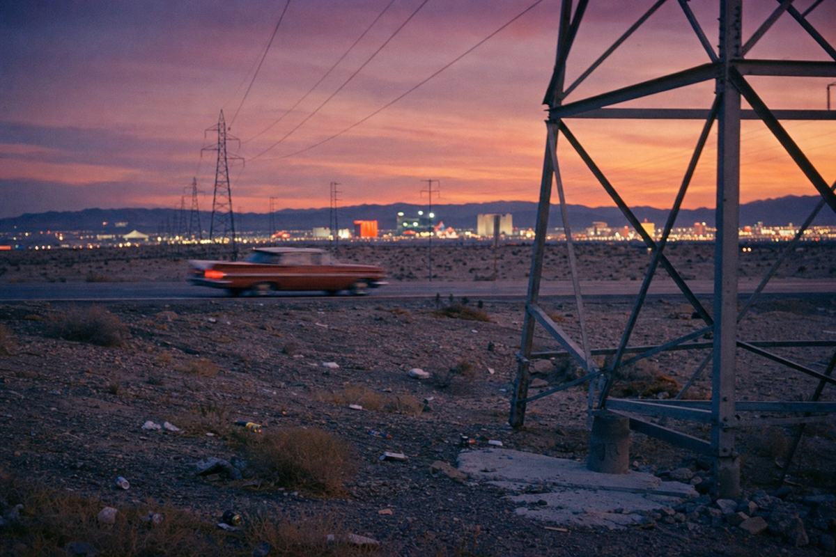 Transmission towers stride in from the desert toward the glow of Las Vegas neon at dusk, late 1950s.