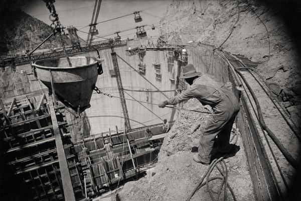 1933 silver gelatin photograph taken from a narrow catwalk on the Nevada rim looking diagonally down across Black Canyon at the rising face of Boulder Dam; cableways and steel derricks crisscross above the concrete; a suspended concrete bucket swings into wooden formwork while a rigger in dust-caked overalls leans into a tag line, his brimmed hat pulled low; additional workers appear as small figures on the dam face; midday desert haze with deep shadows under the rim; film/process: contact print from a 5x7 inch panchromatic negative on fiber-based silver gelatin paper, moderate grain, high contrast, slight edge vignetting, faint handling scratches; lens: approximately 180mm on a view camera; asymmetric composition with the bucket entering from upper left; environment details include hoses, aggregate spill, and rock scree on ledges; no legible text or signage anywhere.