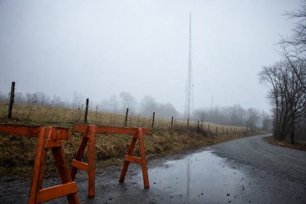 Realistic digital press photograph taken on a cool early morning in Noble County, Ohio: Noble Ridge Mast No. 2 rises from a foggy pasture while two orange wooden road-closure sawhorses with chipped paint sit foreground left on damp gravel; puddles reflect a partial silhouette of the mast. The narrow curve of Noble Ridge Rd SE runs diagonally, with flattened weeds and scattered grit along the verge. The mast is off-center to the right, half-veiled by low clouds; leafless trees and wire fence posts fade into mist. Captured handheld on a modern DSLR with a 50mm lens; shallow depth of field with slight edge falloff; subtle high-ISO noise in the fog; natural cool color balance. No legible text anywhere; asymmetrical, candid composition.